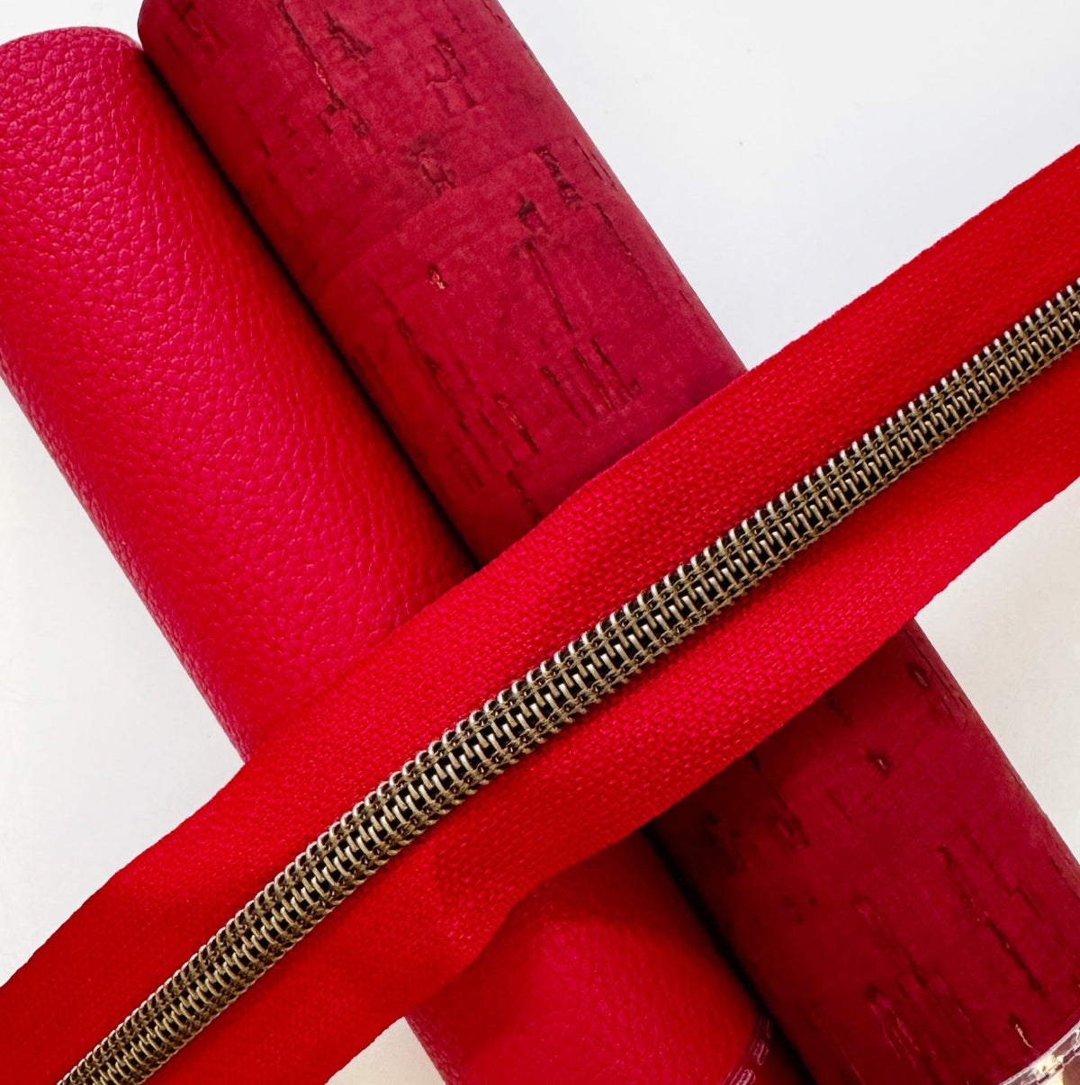 Red continuous zipper tape with bronze teeth lying diagonally across rolls of red faux leather and cork fabric.