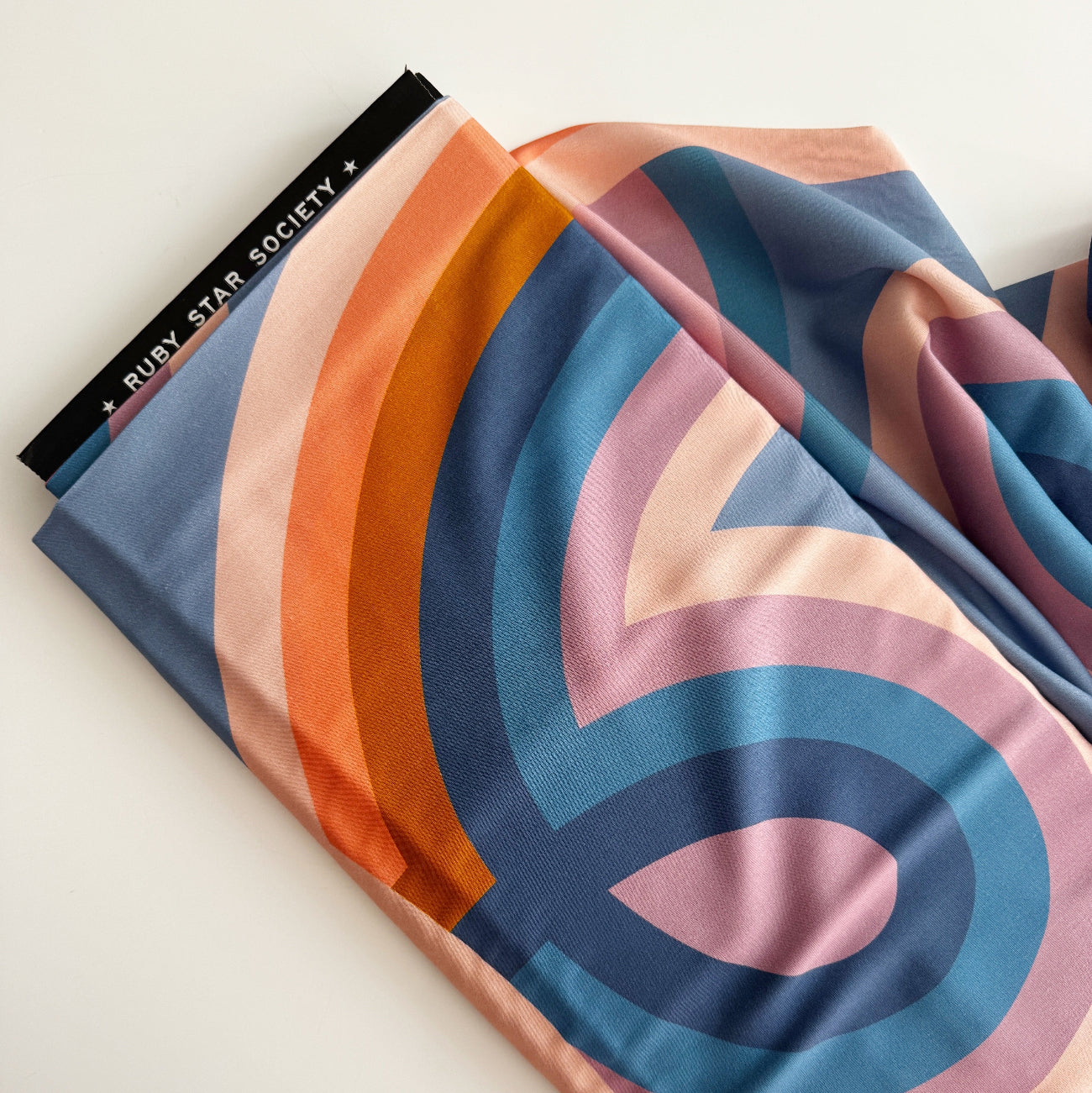 folded length of Ruby Star Society rayon fabric showing a branded black selvage edge and an abstract design of layered, curved bands in blue, peach, orange, and purple.