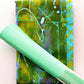 A mint green textured pen resting diagonally across a colorful abstract notebook with green, yellow, and turquoise artwork on a light pink background.