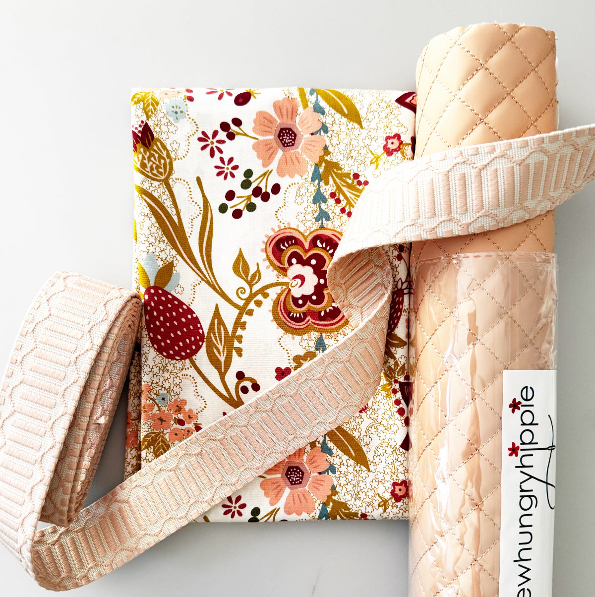 A styled product display featuring a floral fabric yardage with a strawberry pattern, a roll of light peach quilted fabric, and a wide cream lace trim. A white paper label with the brand name 'newhungryhippie' is attached to the roll of fabric, set against a clean white background.