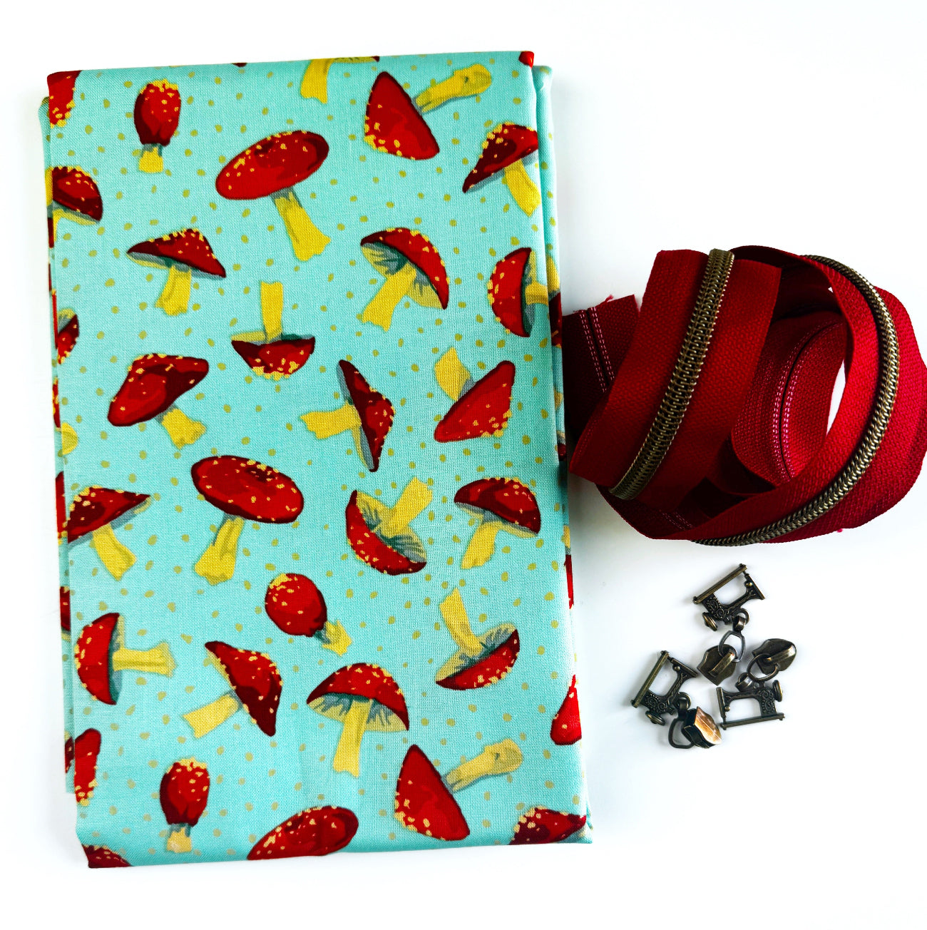 Close-up flat lay of a sewing kit containing teal mushroom-patterned fabric, a red zipper by the yard, and decorative pulls.