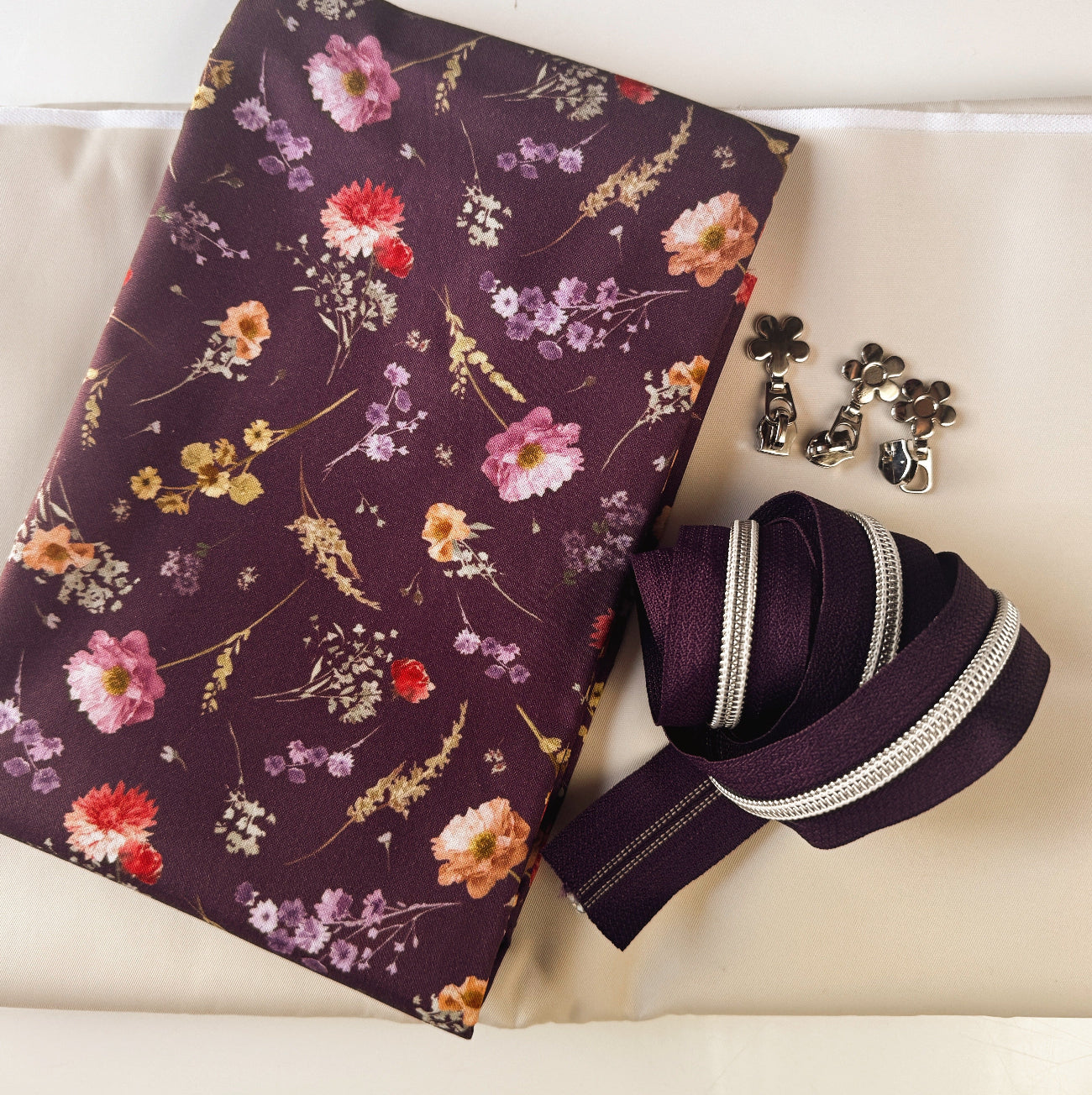 Flat-lay product view of a bag-making kit with floral cotton fabric, cream bonded nylon lining, plum zipper tape, and decorative flower pulls.