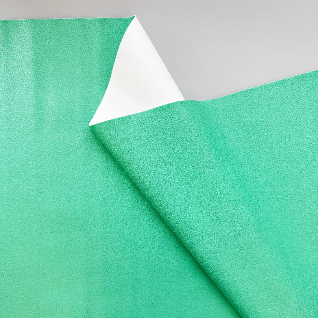 A close-up of a mint green textured sheet, likely vinyl or adhesive film, with a folded corner revealing a white backing, set against a neutral light gray background.