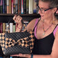 A woman wearing glasses and a black top with pink straps holds up a black quilted handbag with a beige and black checkered bow detail, set against a background of bookshelves.