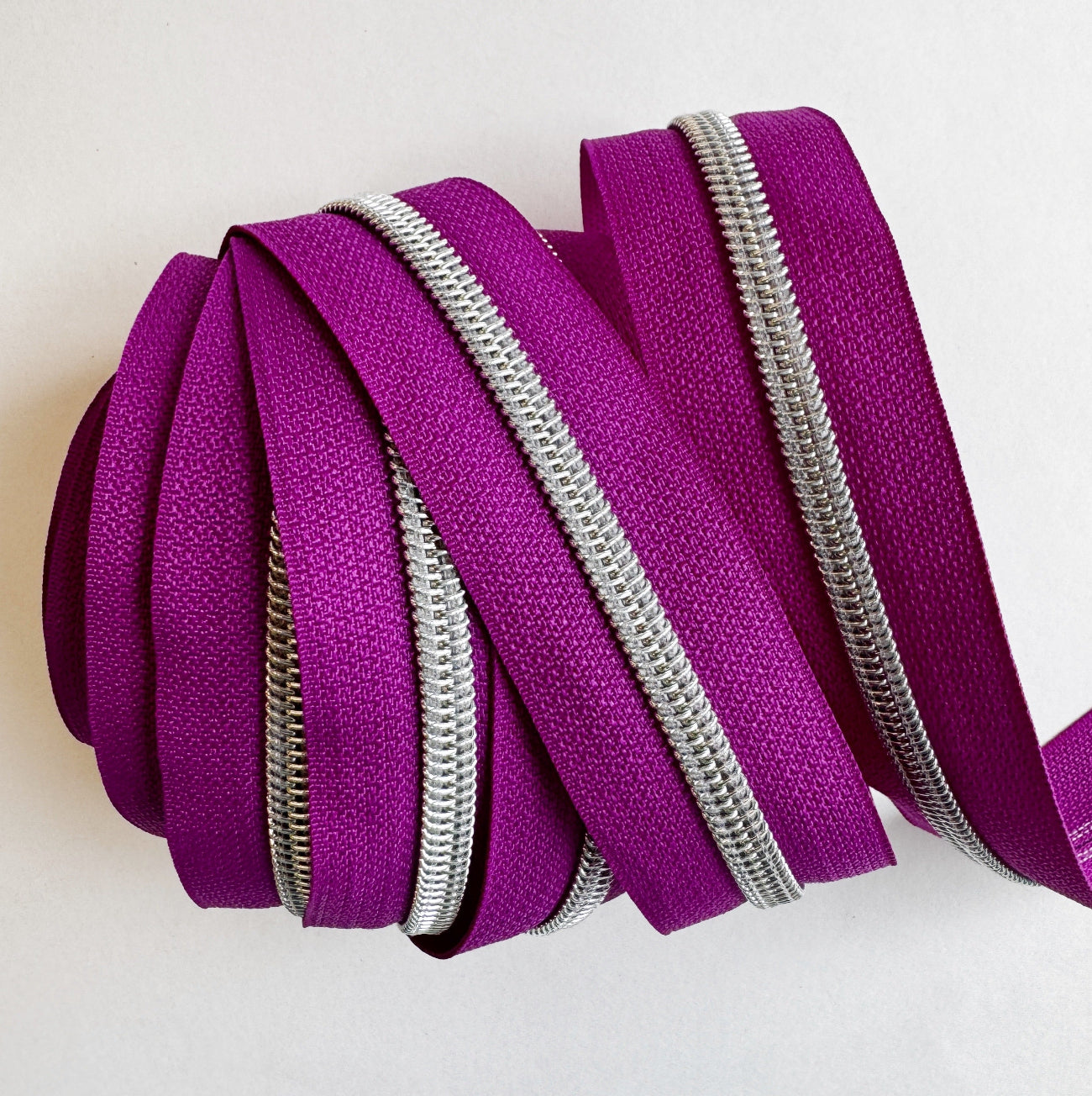 A close-up view of bright teal nylon zipper tape neatly layered and wrapped flatly to showcase several vertical, parallel rows of shiny silver metallic coil teeth against a plain white background.