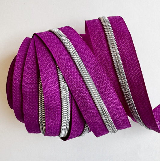 A close-up view of bright teal nylon zipper tape neatly layered and wrapped flatly to showcase several vertical, parallel rows of shiny silver metallic coil teeth against a plain white background.