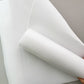 A close-up of a white sheet of material being held back by a hand, showing its smooth, slightly translucent texture against a neutral light gray background.