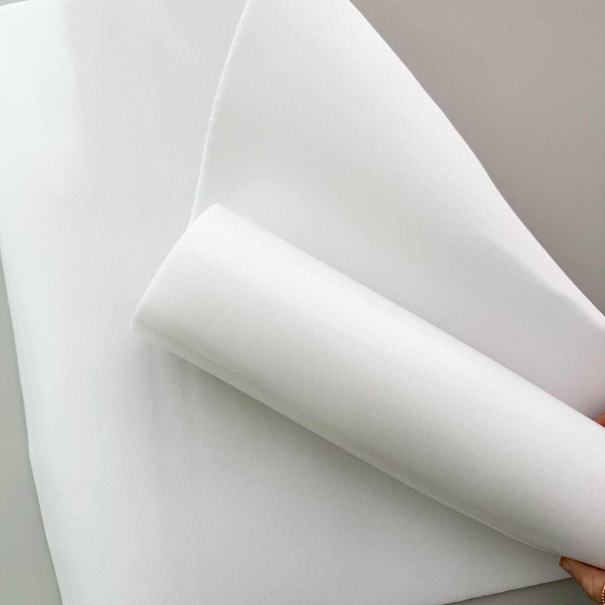 A close-up of a white sheet of material being held back by a hand, showing its smooth, slightly translucent texture against a neutral light gray background.
