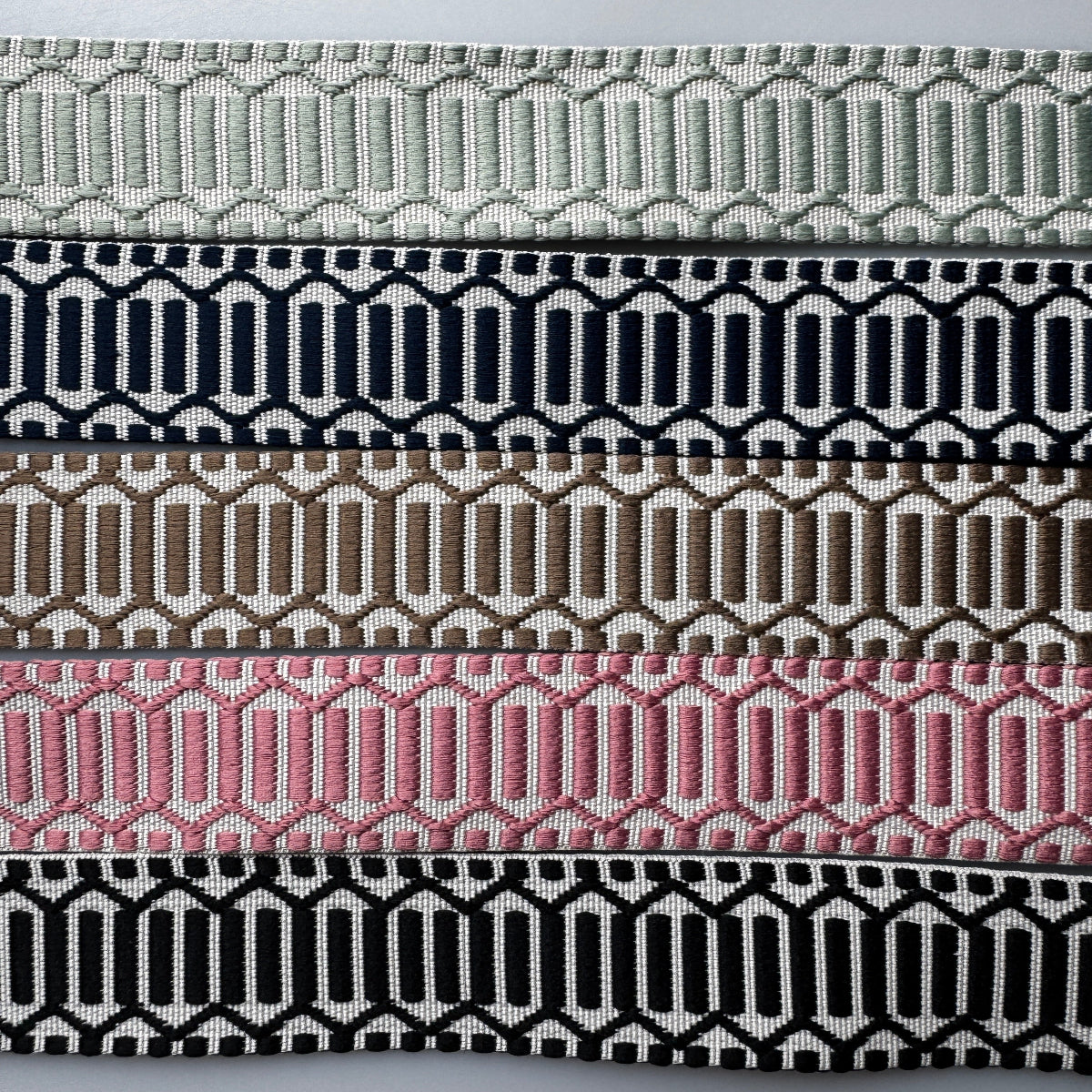 Five horizontal woven trim or ribbon strips with geometric patterns in different colors, displayed on a light gray background. The colors are light sage green, navy blue, taupe brown, pink, and black.