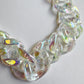 A close-up, top-down view of a thick acrylic curb chain links strand laid out on a plain white surface. The chain links are made of a clear, translucent material with a glossy, iridescent finish that catches the light, reflecting soft pastel shades of yellow, pink, purple, and green.