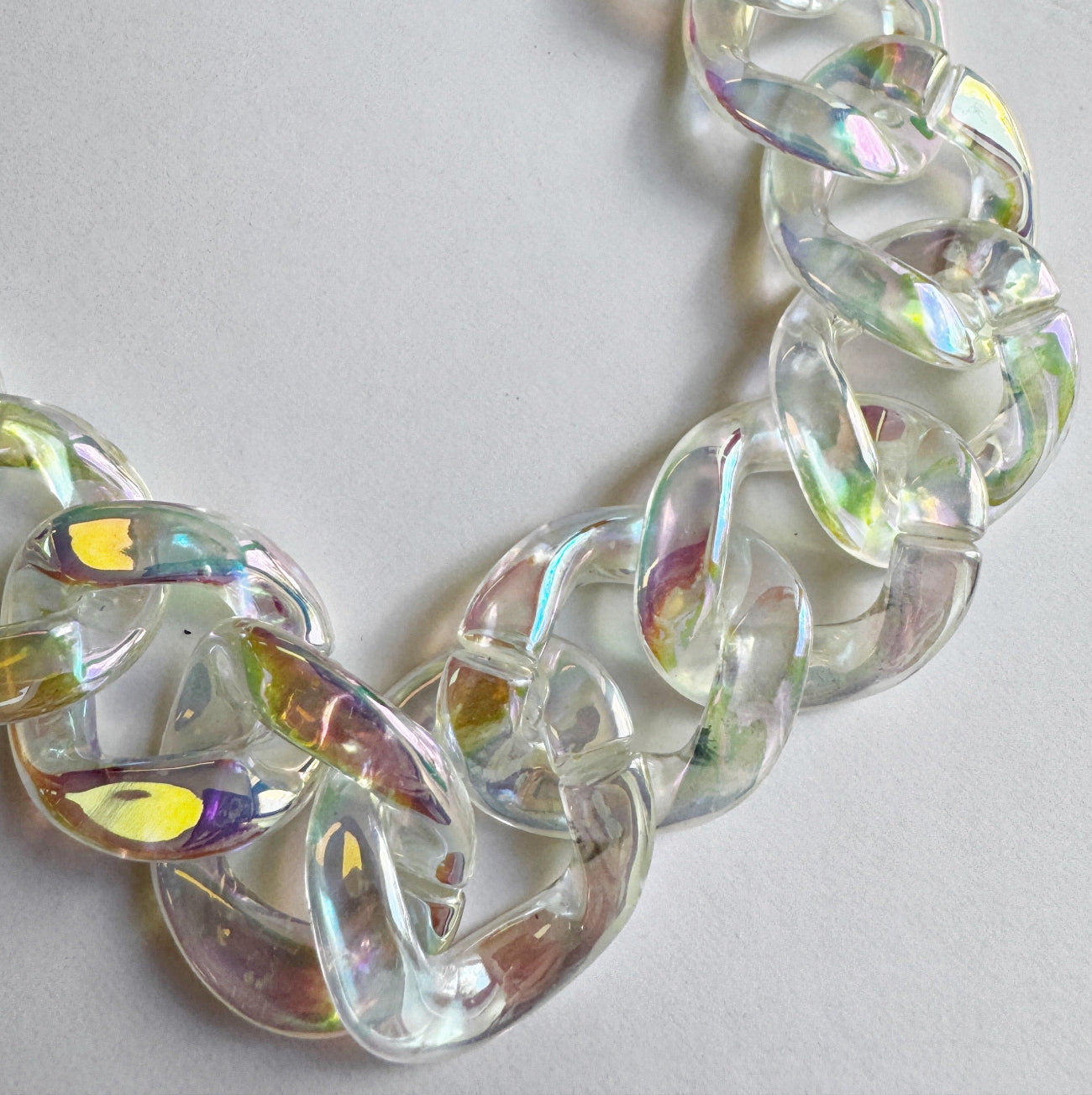 A close-up, top-down view of a thick acrylic curb chain links strand laid out on a plain white surface. The chain links are made of a clear, translucent material with a glossy, iridescent finish that catches the light, reflecting soft pastel shades of yellow, pink, purple, and green.