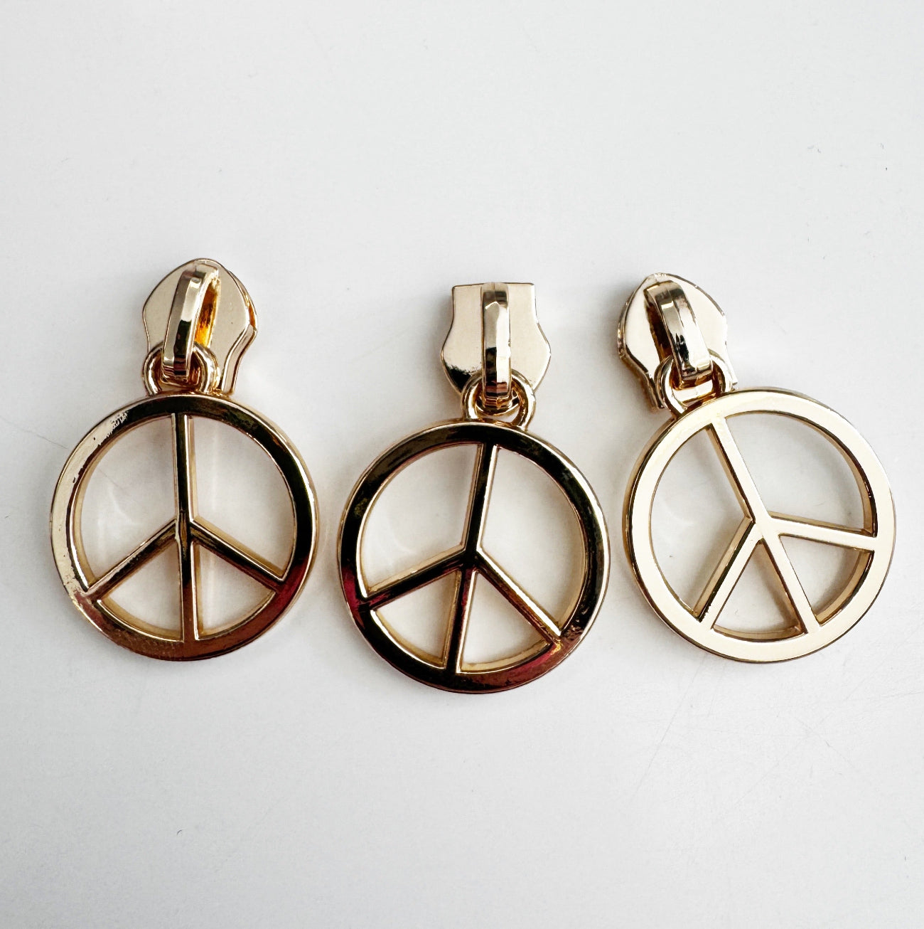 Close-up of three identical gold-colored zipper slider bodies attached to circular peace symbol charms, resting on a flat white surface.
