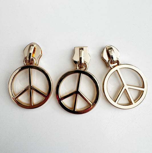 Close-up of three identical gold-colored zipper slider bodies attached to circular peace symbol charms, resting on a flat white surface.