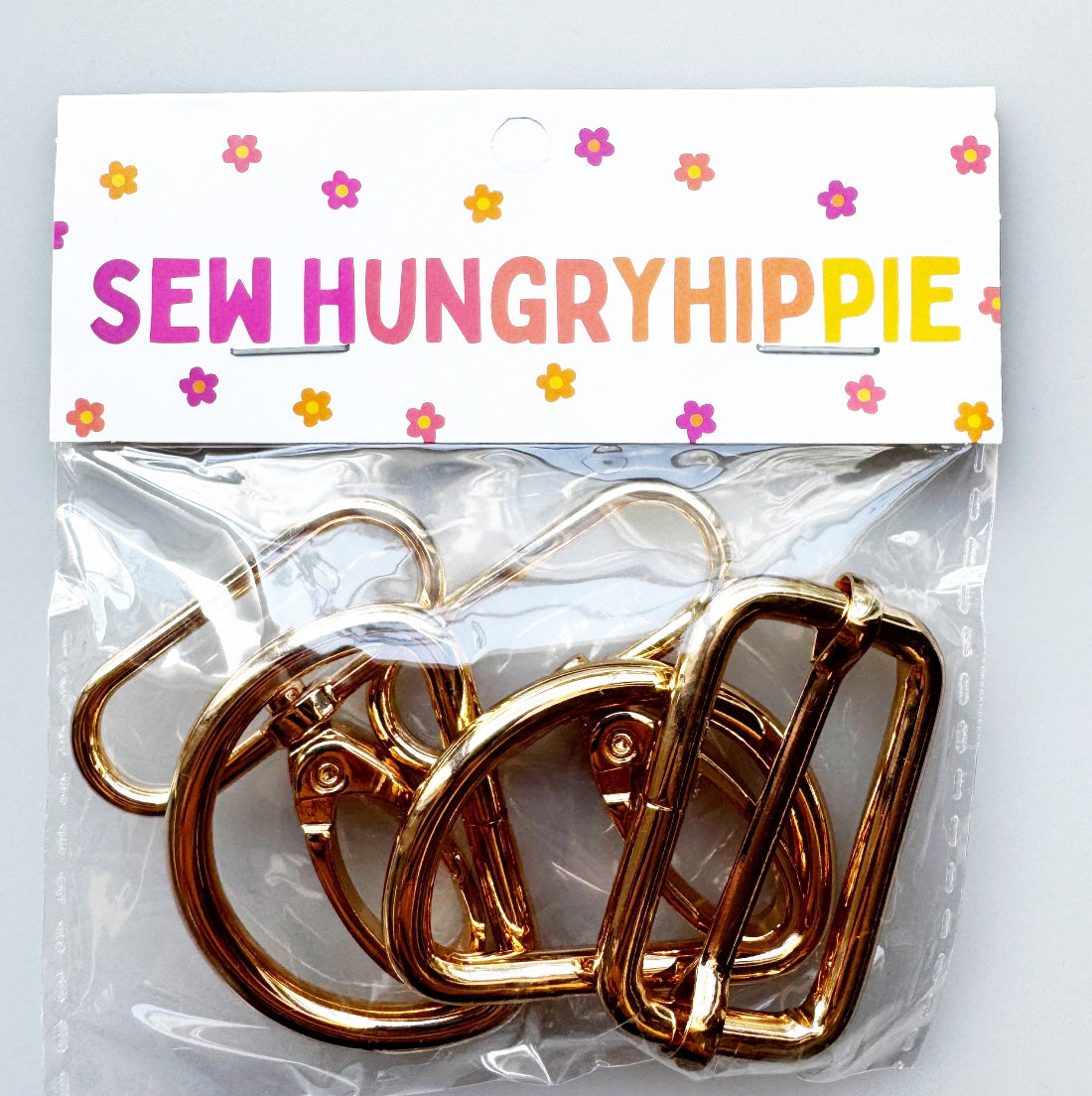 Three  gold metal sewing clips inside a clear plastic bag with a white header card that reads 'SEW HUNGRYHIPPIE' in colorful letters and has small pink, purple, and orange flowers.