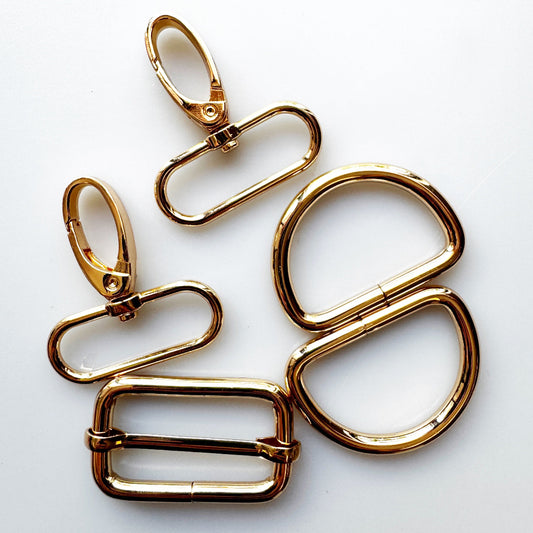 Five gold-toned metal keychain or bag hardware pieces, including two lobster clasp rings, two D-rings, and one double-buckle bar, arranged on a plain white background.