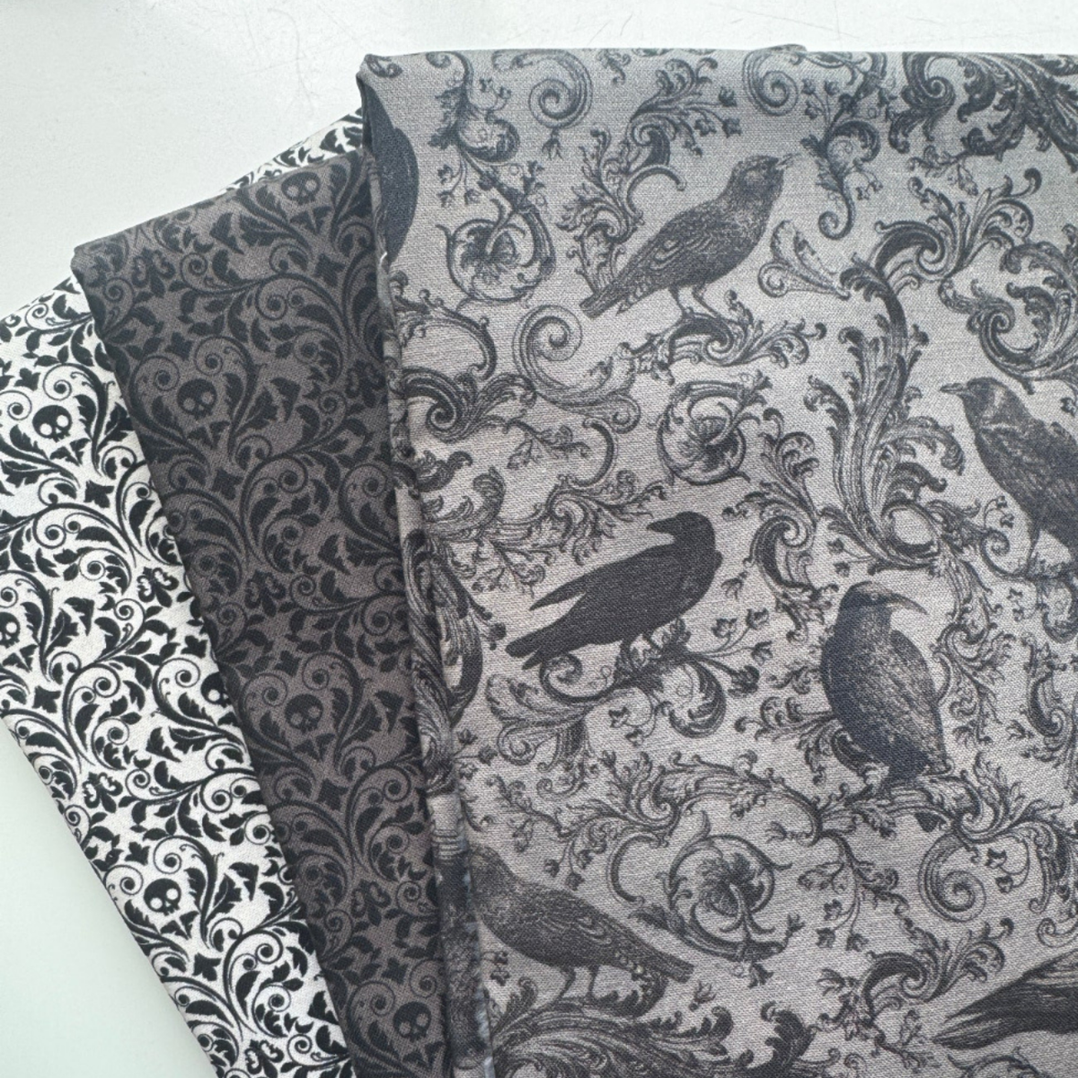 Overhead, close-up shot of three distinct cotton quilting fabrics fanned out and overlapping gothic, monochromatic color scheme of black, white, and shades of gray. The topmost fabric is a medium gray tone, printed with highly detailed illustrations of perched ravens woven into a complex, swirling vintage damask scroll pattern. Partially tucked underneath is a dark charcoal fabric featuring a subtle, dense damask design seamlessly hiding small skulls. 