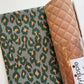 Bag-making fabric set featuring green-gray cotton fabric with a forest green and peach-tan abstract ikat leopard print, paired with camel-brown diamond-quilted faux leather vinyl in protective plastic wrapping. Coordinating cotton quilting fabric and quilted vinyl for handmade handbags, purses, tote bags, and DIY sewing projects.