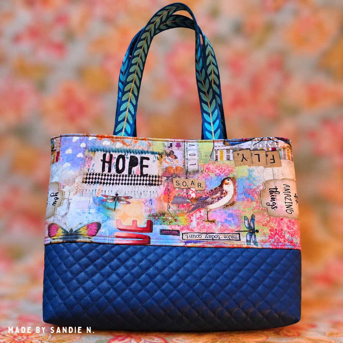Handmade fabric tote bag featuring a colorful bird and dragonfly collage print with inspirational words like HOPE and SOAR, a dark navy blue diamond-quilted vinyl base, and blue-green leaf-patterned ribbon shoulder straps. Bag handmade by Sandie N.