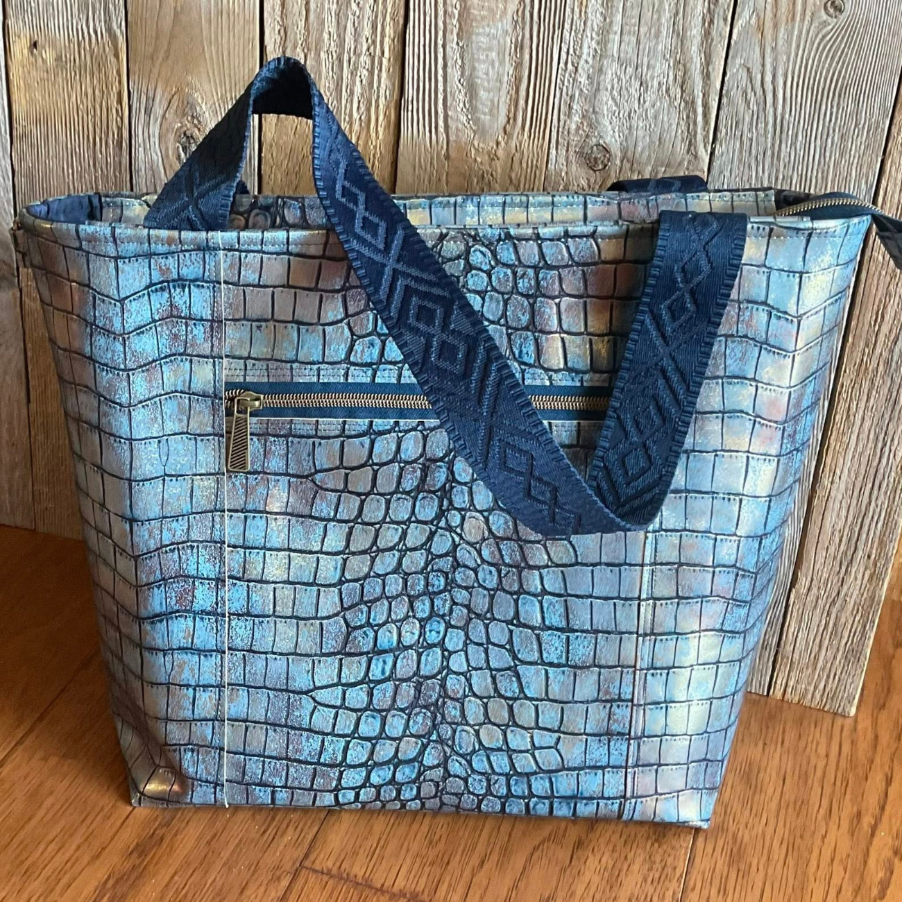 Handmade zipper-top Santorini tote bag crafted from iridescent blue and silver crocodile embossed faux leather, featuring an exterior zipper pocket and navy blue geometric diamond jacquard webbing handles.