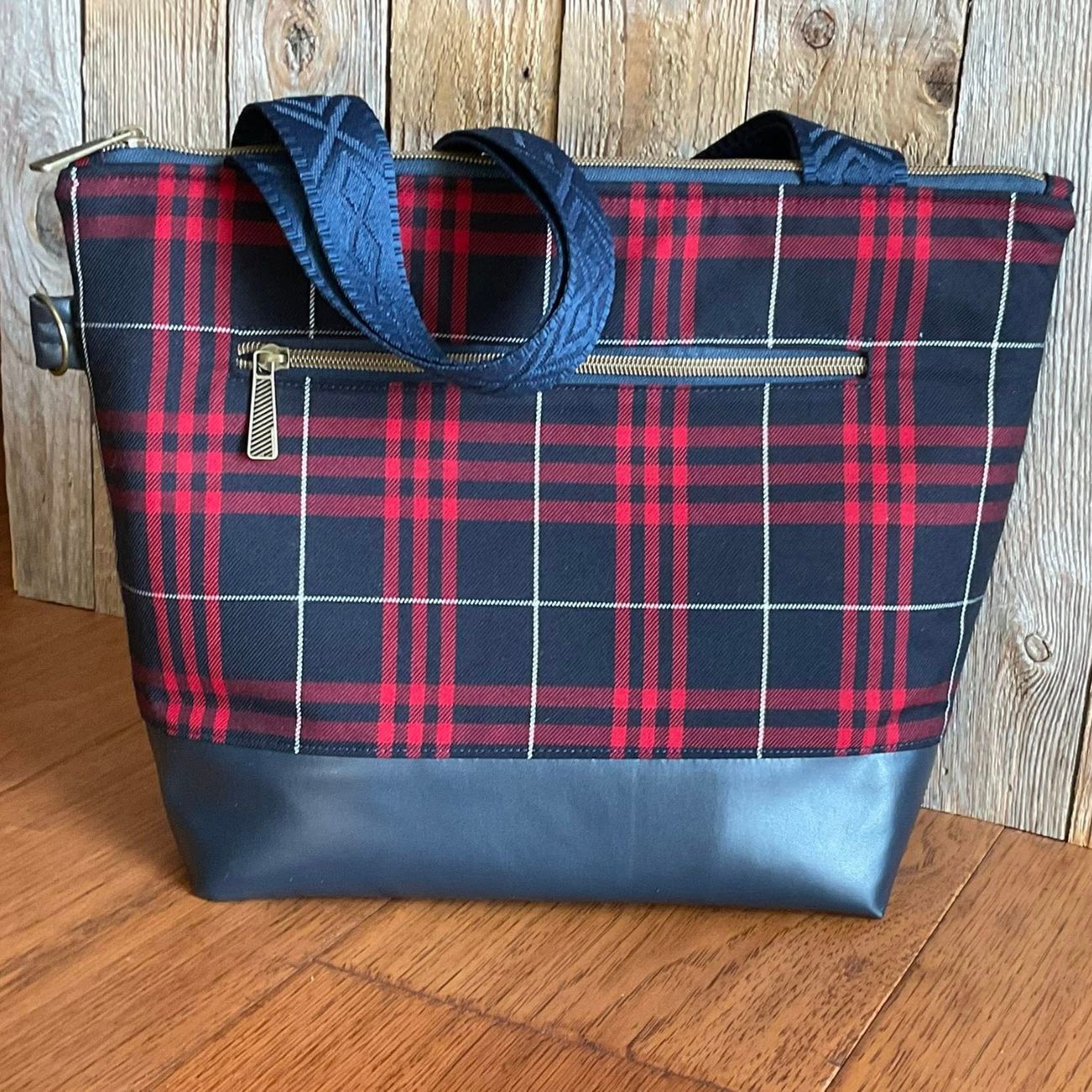 Handmade zipper top tote bag featuring a red, black, and white plaid fabric body, navy vinyl base, brass exterior zipper pocket, and blue geometric diamond jacquard webbing straps.