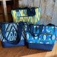 Three handmade zipper-top tote bags featuring blue faux leather bases, zippered exterior pockets, and navy blue geometric diamond jacquard webbing straps, shown in yellow ikat, black ikat, and blue abstract swirl prints.