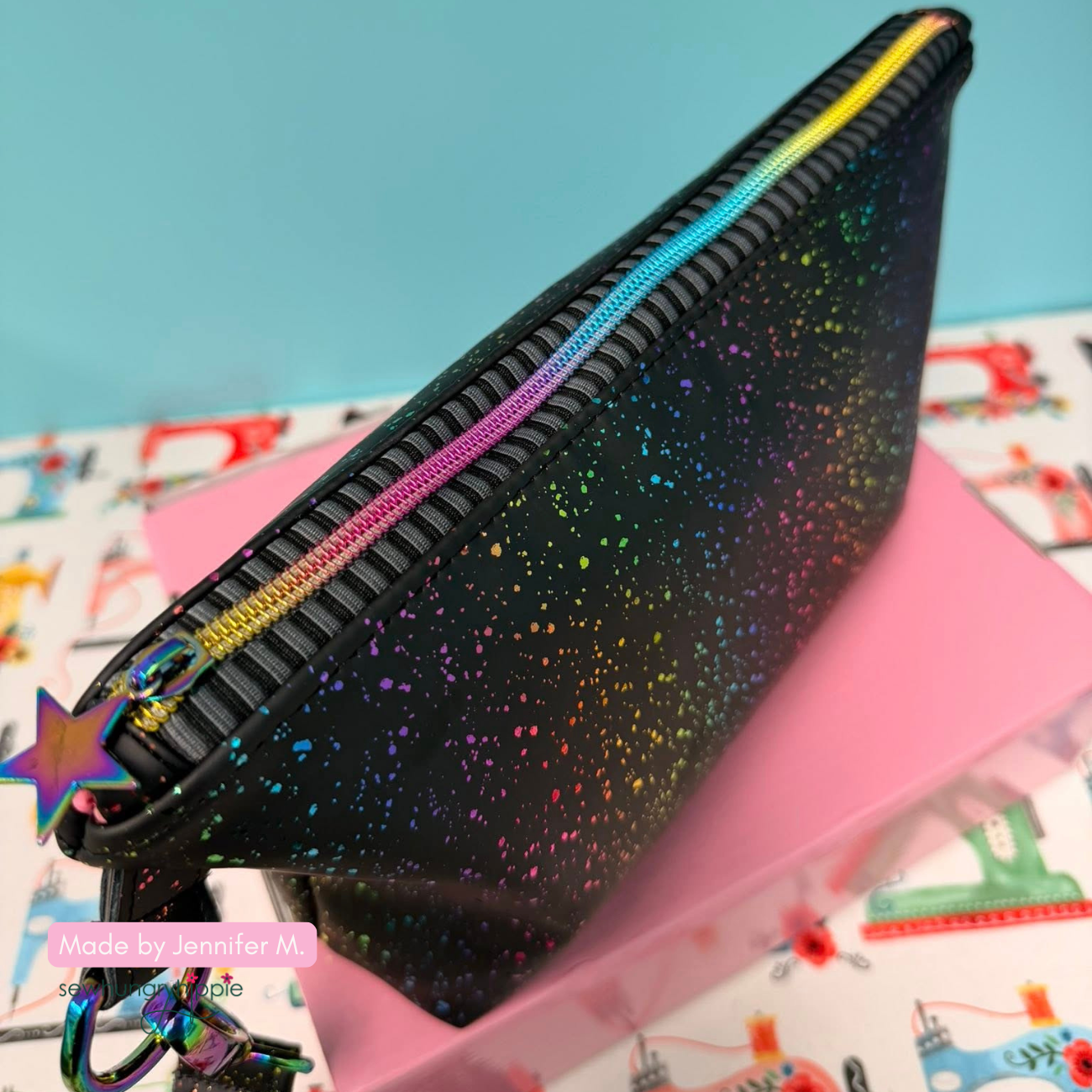A finished handmade zipper pouch made with black vinyl featuring a rainbow speckled print, constructed using a charcoal and black striped jacquard zipper tape, a rainbow iridescent coil track, and a matching star-shaped zipper pull.