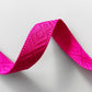 A close-up studio shot of a wavy section of vibrant hot pink jacquard woven webbing strap showing an intricate diamond geometric pattern on a white background