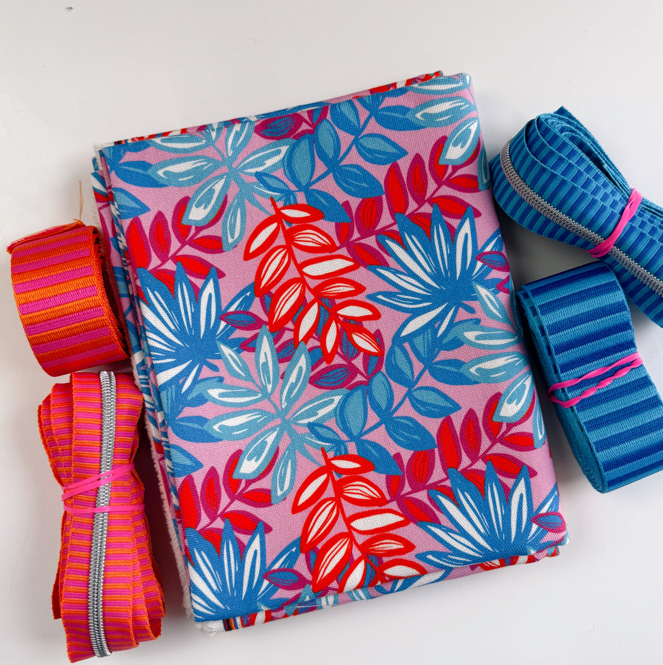 uly Canvas Club bundle featuring a folded pink and blue tropical leaf print canvas fabric surrounded by coordinating striped orange and blue bag webbing and zippers by the yard