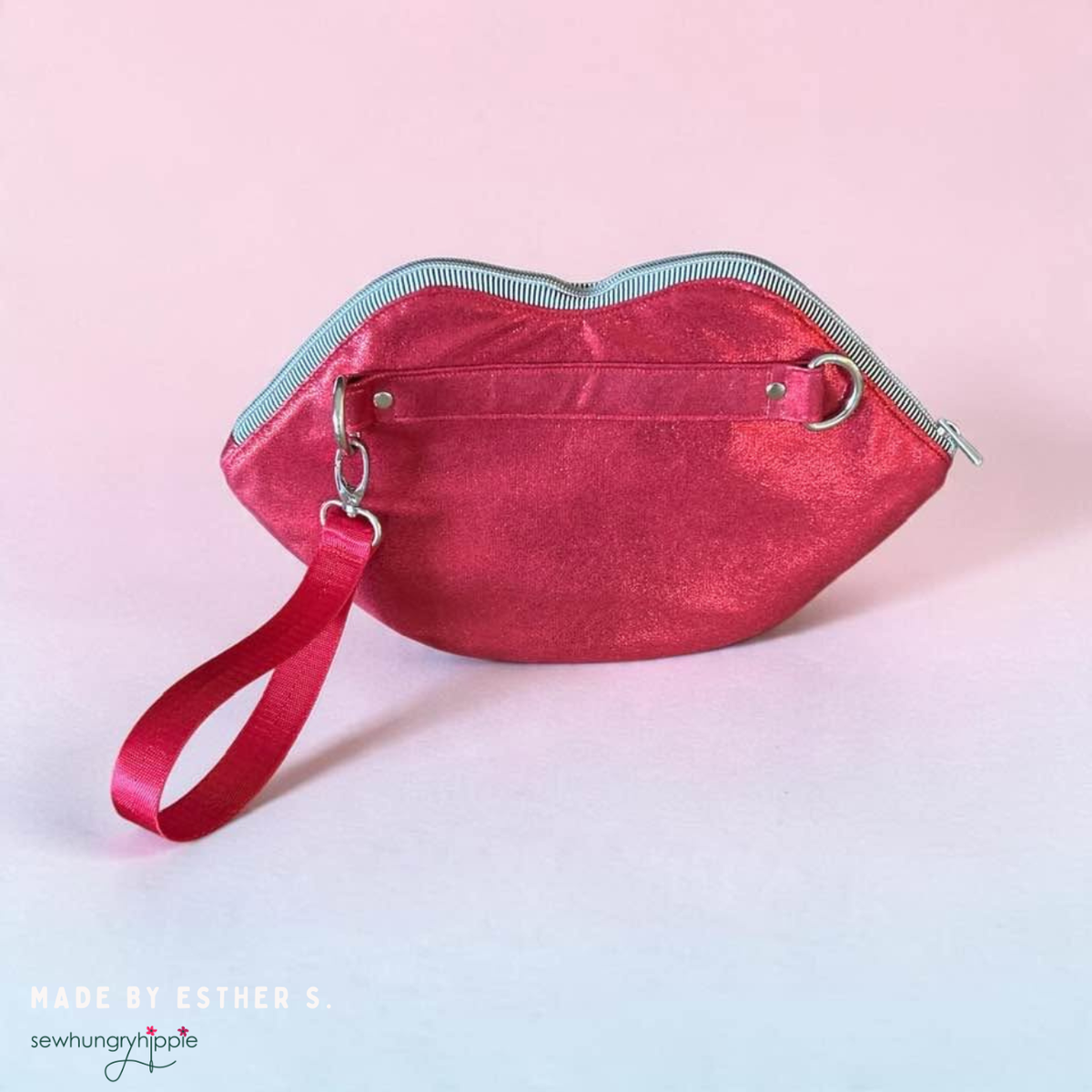 Metallic pinkish-red lips-shaped zipper pouch with a glossy finish, black and white striped zipper trim and matching wristlet strap attached to a metal D-ring, displayed against a light pink background. Unique handmade lip-shaped coin purse, cosmetic pouch, or statement bag accessory.