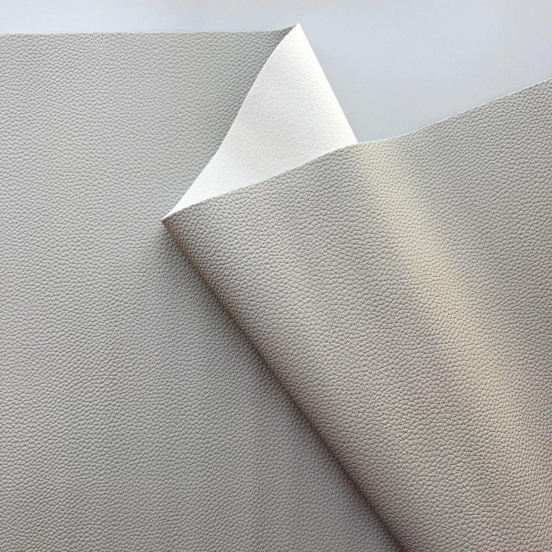 A close-up of a light gray pebbled material sheet with one corner folded back to show the lighter underside, photographed on a plain light background.