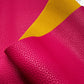 A swatch of double-sided pebble-grained leather fabric showing a vibrant fuchsia pink exterior and a contrasting mustard yellow backing material