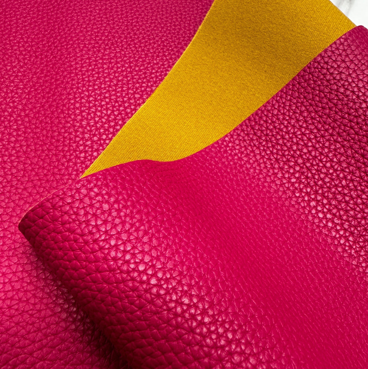 A swatch of double-sided pebble-grained leather fabric showing a vibrant fuchsia pink exterior and a contrasting mustard yellow backing material