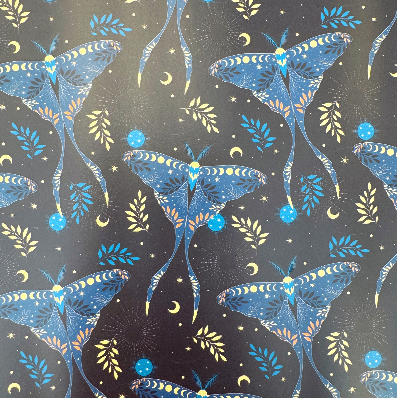 A seamless repeating pattern in the file luna moth vinyl roll 18 x 54.jpg featuring large, stylized blue luna moths on a dark charcoal-black background. The moths have intricate wing details showing moon phases and botanical leaves. The background is filled with scattered celestial elements like yellow crescent moons, tiny stars, faint sunbursts, glowing blue orbs, and small leafy sprigs in blue and yellow.