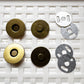 Six circular metal magnetic button components arranged on a white textured foam background, including two brass-toned pieces, two dark bronze-toned pieces, and two silver-toned metal washers.