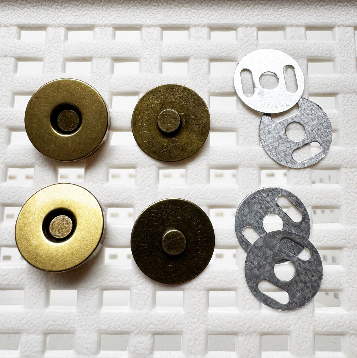 Six circular metal magnetic button components arranged on a white textured foam background, including two brass-toned pieces, two dark bronze-toned pieces, and two silver-toned metal washers.