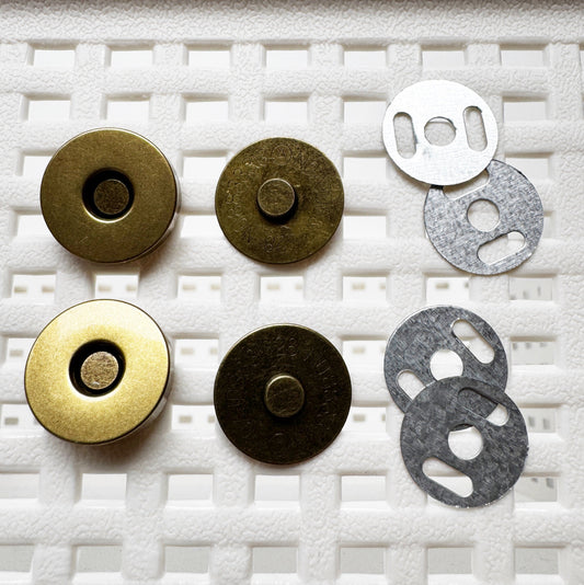 Six circular metal magnetic button components arranged on a white textured foam background, including two brass-toned pieces, two dark bronze-toned pieces, and two silver-toned metal washers.
