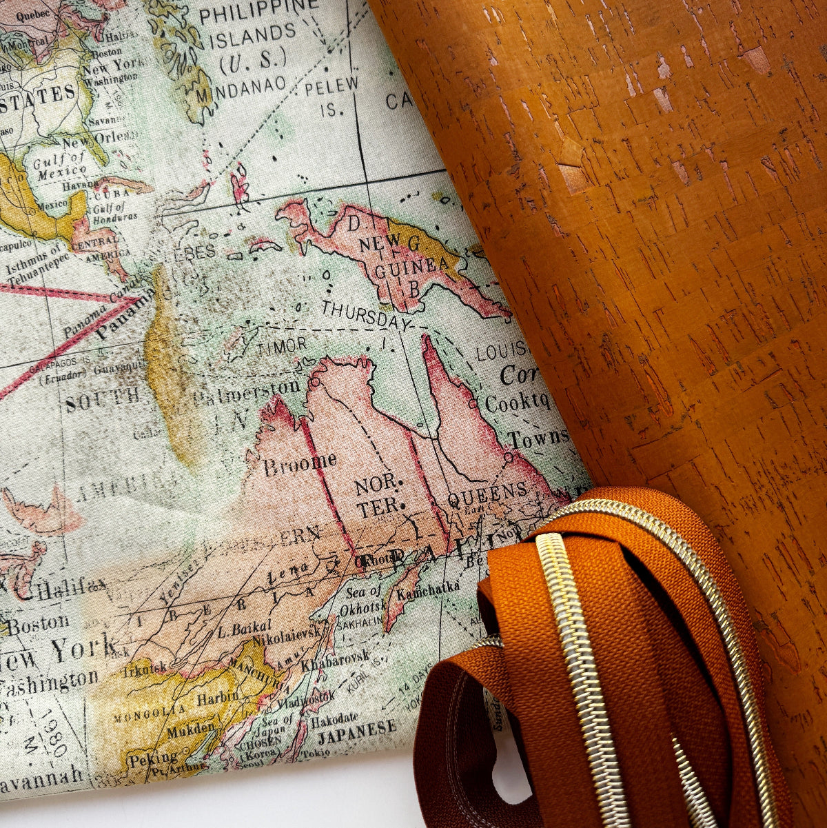 A close-up of a vintage-style map featuring North America with soft pastel country colors, partially rolled over to reveal a textured orange-brown cork-like underside. A matching rolled strip with a gold-tone zipper runs across the lower right corner.