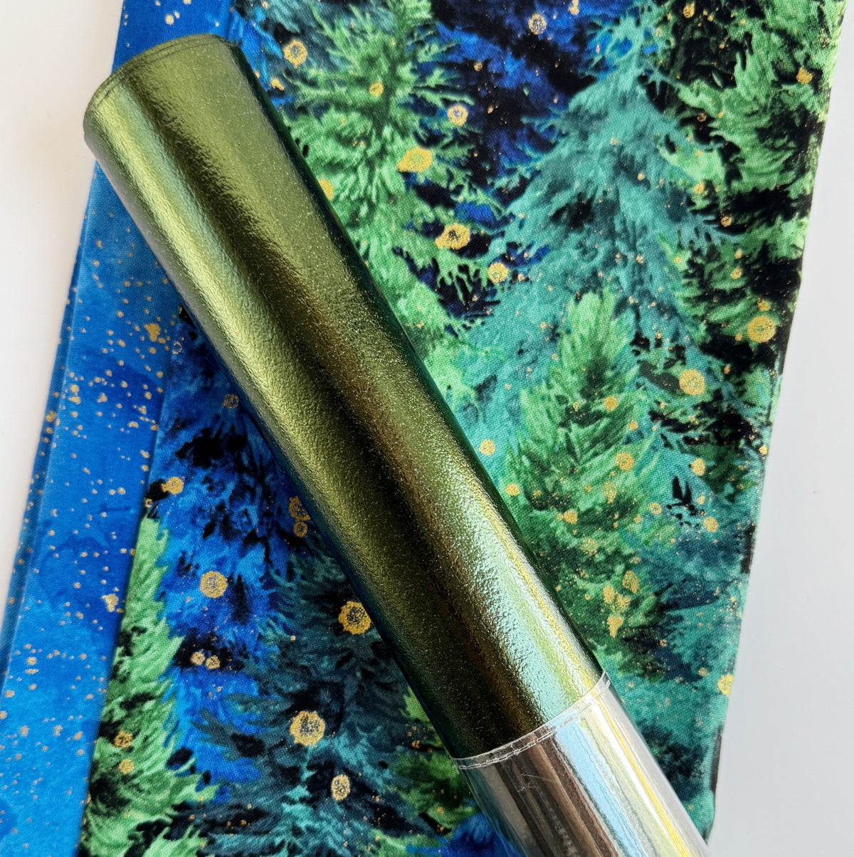 A roll of metallic green vinyl fabric resting on top of patterned blue and green forest-themed fabric.