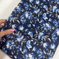 A close-up, angled view of the Midnight Bloom - Navy Blue Bonded Nylon – 36" x 58" fabric being held by a hand with pink manicured nails and a gold ring. The navy blue bonded nylon fabric features a striking floral pattern with shades of light blue and white. The shot captures the smooth texture and slight sheen of the material as it folds naturally under the hand's grip.
