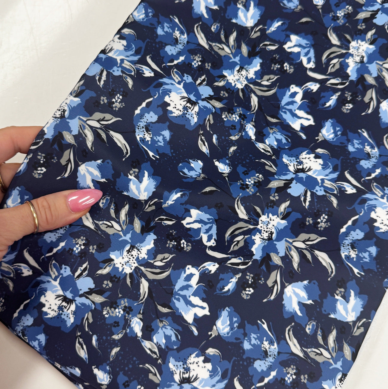 A close-up, angled view of the Midnight Bloom - Navy Blue Bonded Nylon – 36" x 58" fabric being held by a hand with pink manicured nails and a gold ring. The navy blue bonded nylon fabric features a striking floral pattern with shades of light blue and white. The shot captures the smooth texture and slight sheen of the material as it folds naturally under the hand's grip.