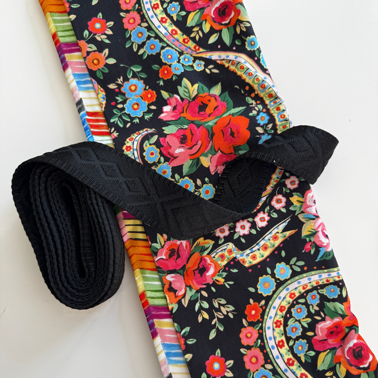 A sewing bundle featuring a folded vibrant floral fabric with red roses, blue blossoms, and multi-colored paisley borders on a black background, paired with a bundled roll of black jacquard woven webbing strap displaying a raised diamond geometric pattern.