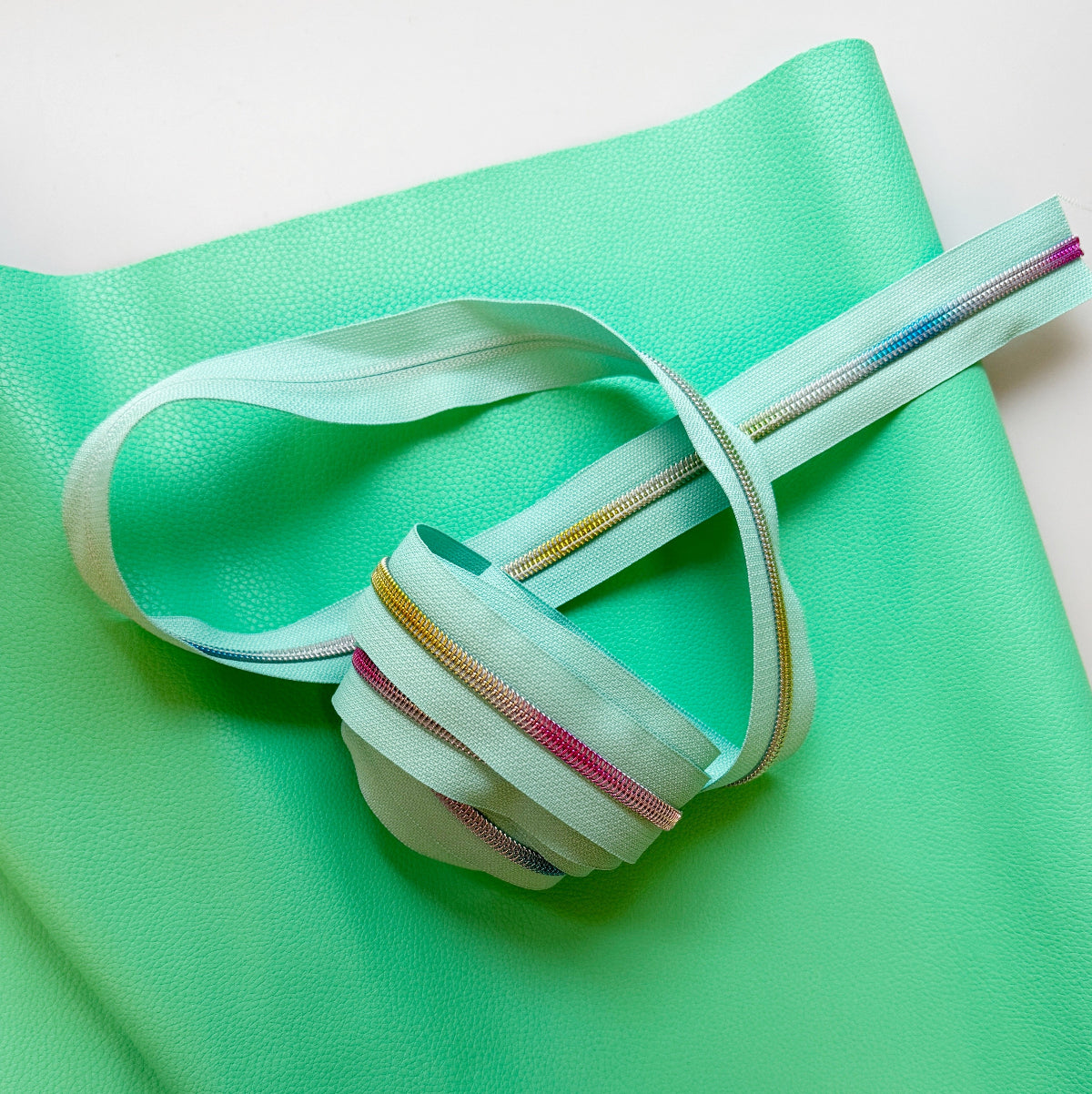 A mint green zipper with thin metallic silver, gold, pink, and blue stripes, wrapped and draped over a matching mint green textured material on a white background.