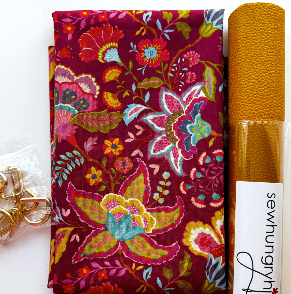 Moda Fabrics Sitara Jasmine in Rose Wine cotton quilting fabric by Crystal Manning featuring vibrant Bohemian floral and paisley motifs in fuchsia, teal, lime green, mustard yellow, orange, and cream on a burgundy background, paired with mustard yellow textured faux leather vinyl and gold-tone lobster clasps and D-rings from Sew Hungry Hippie. Coordinated bag-making supplies for DIY handbags, purses, and sewing projects.