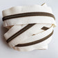 A coiled bundle of off-white canvas zipper tape with dark bronze metal zippers, photographed on a clean white background.