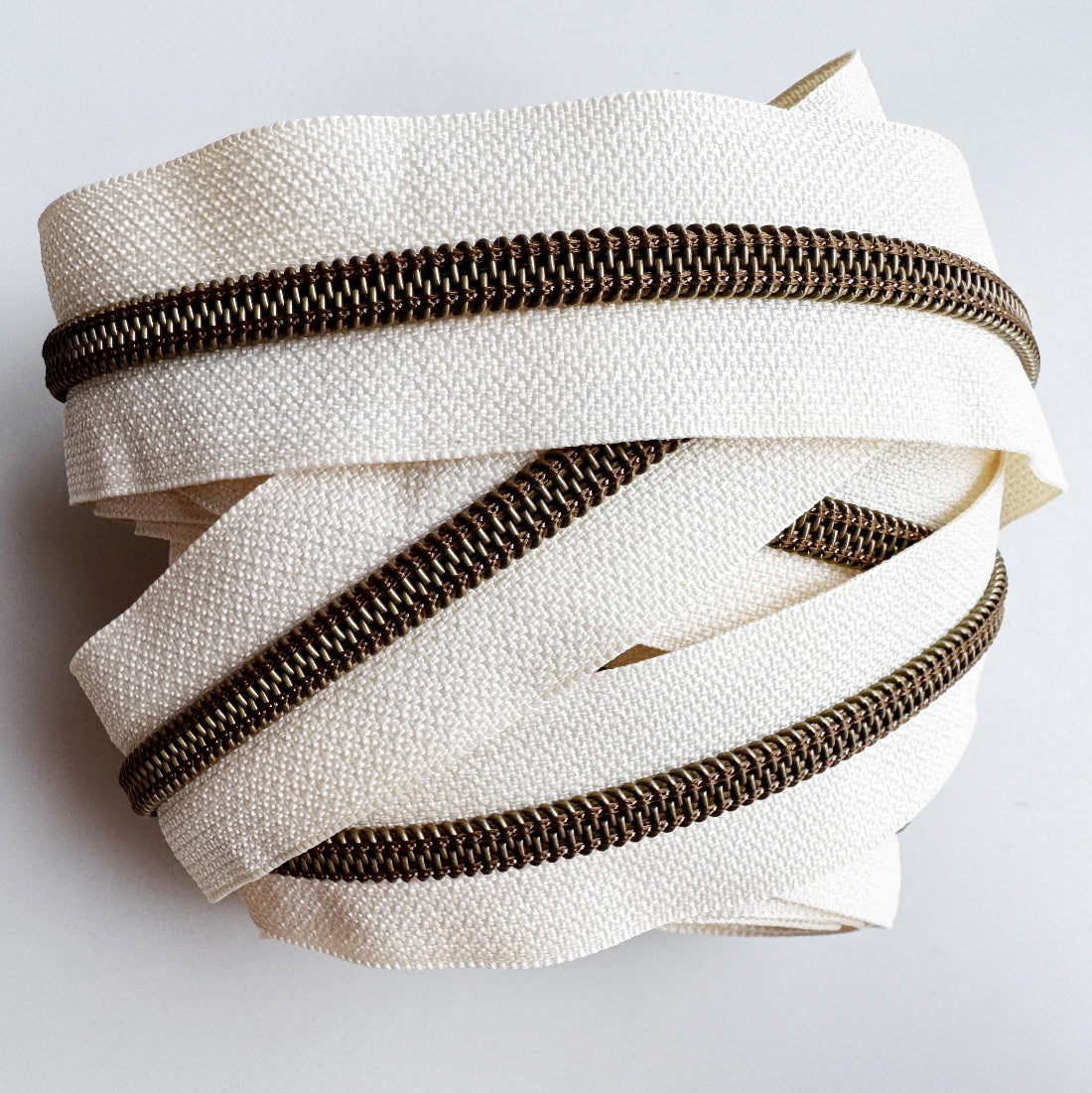 A coiled bundle of off-white canvas zipper tape with dark bronze metal zippers, photographed on a clean white background.