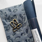 A bag-making material coordination bundle from Sew HungryHippie. The flat lay includes a piece of printed cotton fabric featuring a gray and navy blue botanical leaf pattern, paired with a rolled cylinder of navy blue vegan faux leather fabric. Placed on top of the printed fabric are antique brass bag hardware components, including two swivel snap hooks and a slider buckle