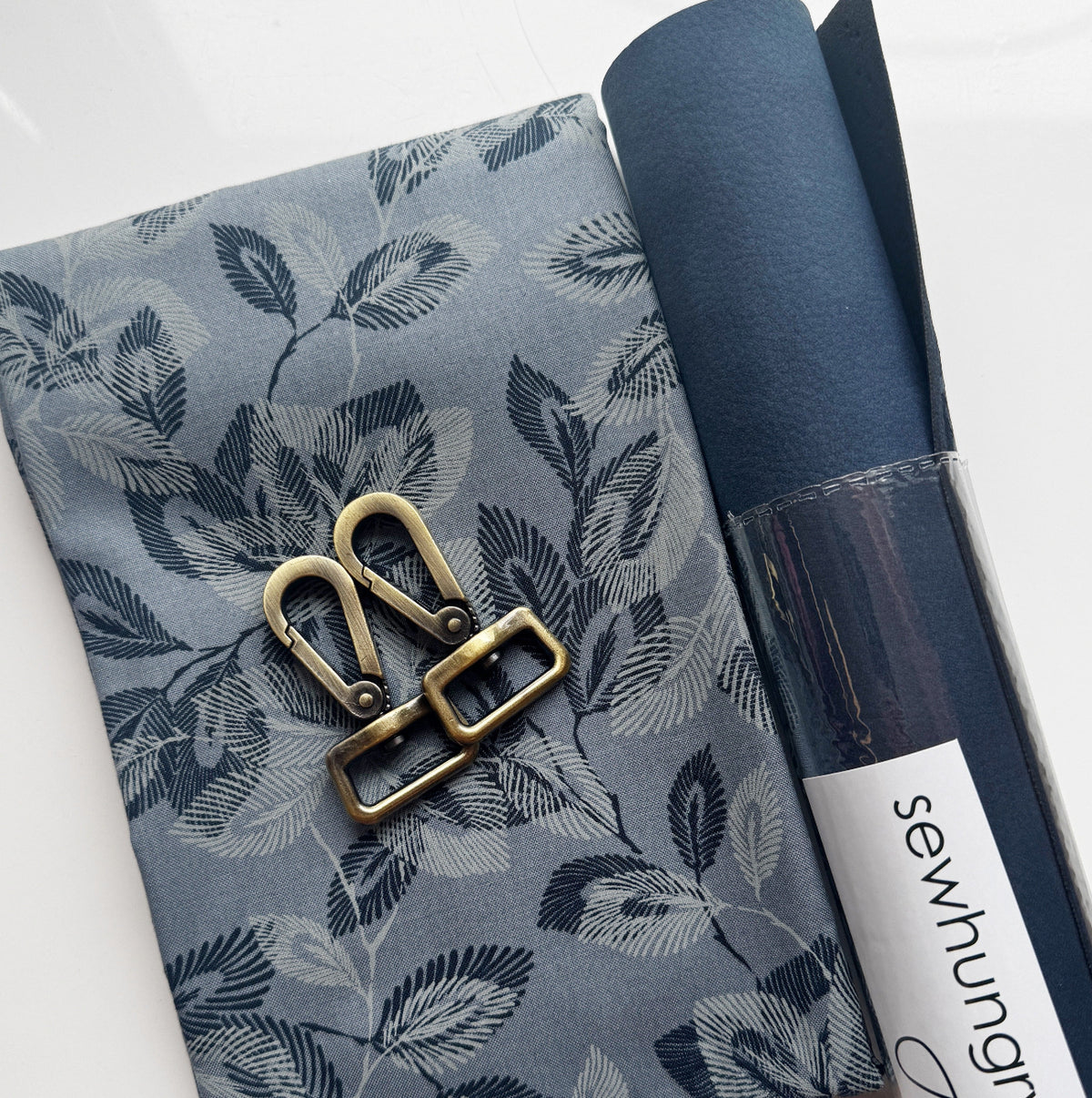 A bag-making material coordination bundle from Sew HungryHippie. The flat lay includes a piece of printed cotton fabric featuring a gray and navy blue botanical leaf pattern, paired with a rolled cylinder of navy blue vegan faux leather fabric. Placed on top of the printed fabric are antique brass bag hardware components, including two swivel snap hooks and a slider buckle