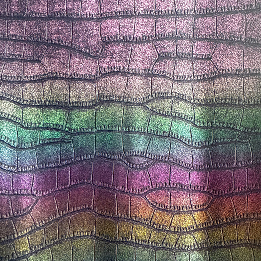 A close-up of a textured material with an embossed reptile or scale pattern in a metallic iridescent finish, showing a gradient of purple, green, and gold tones.