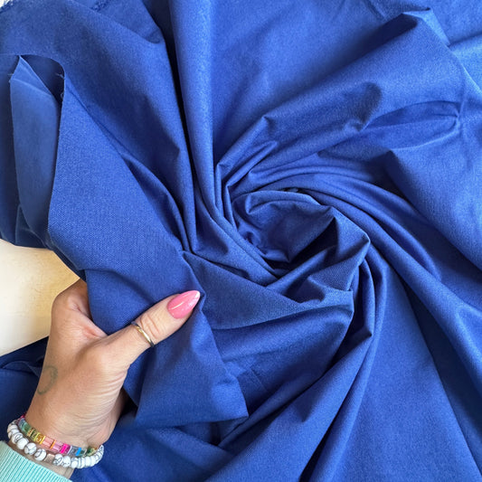 A top-down view of a vibrant royal blue nylon bag lining material, swirled into a spiral pattern with deep folds. In the lower-left corner, a hand with a pink manicure, a gold ring, and colorful beaded bracelets grips a section of the textured, durable fabric to showcase its weight and flexibility.