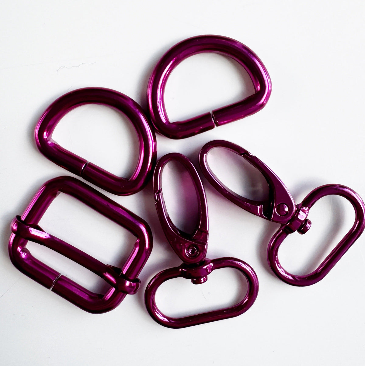 A set of metallic pink or magenta bag-making hardware, including swivel snap hooks, D-rings, and a strap slider buckle on white background